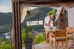 Romantic couple kissing under a pergola with a Marry Me sign, overlooking a scenic hillside view.