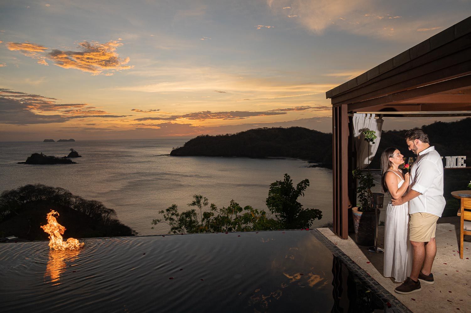 Couple sharing a romantic sunset moment by an infinity pool with ocean views and a fire feature at a tropical resort.