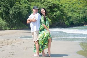 Couple walking barefoot on a tropical beach, woman in green floral dress, man in white shirt and hat.