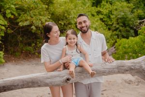 Happy family of three with young daughter sitting on driftwood log at tropical beach surrounded by greenery.