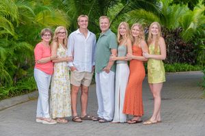 Family of six posing together outdoors in tropical setting, wearing colorful summer outfits among lush green palms.