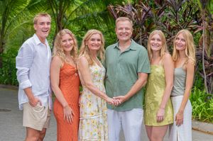A smiling family of six poses together outdoors in a tropical garden setting, dressed in casual summer attire.