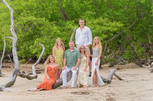 Family of six posing together on a tropical beach surrounded by driftwood and lush green trees.