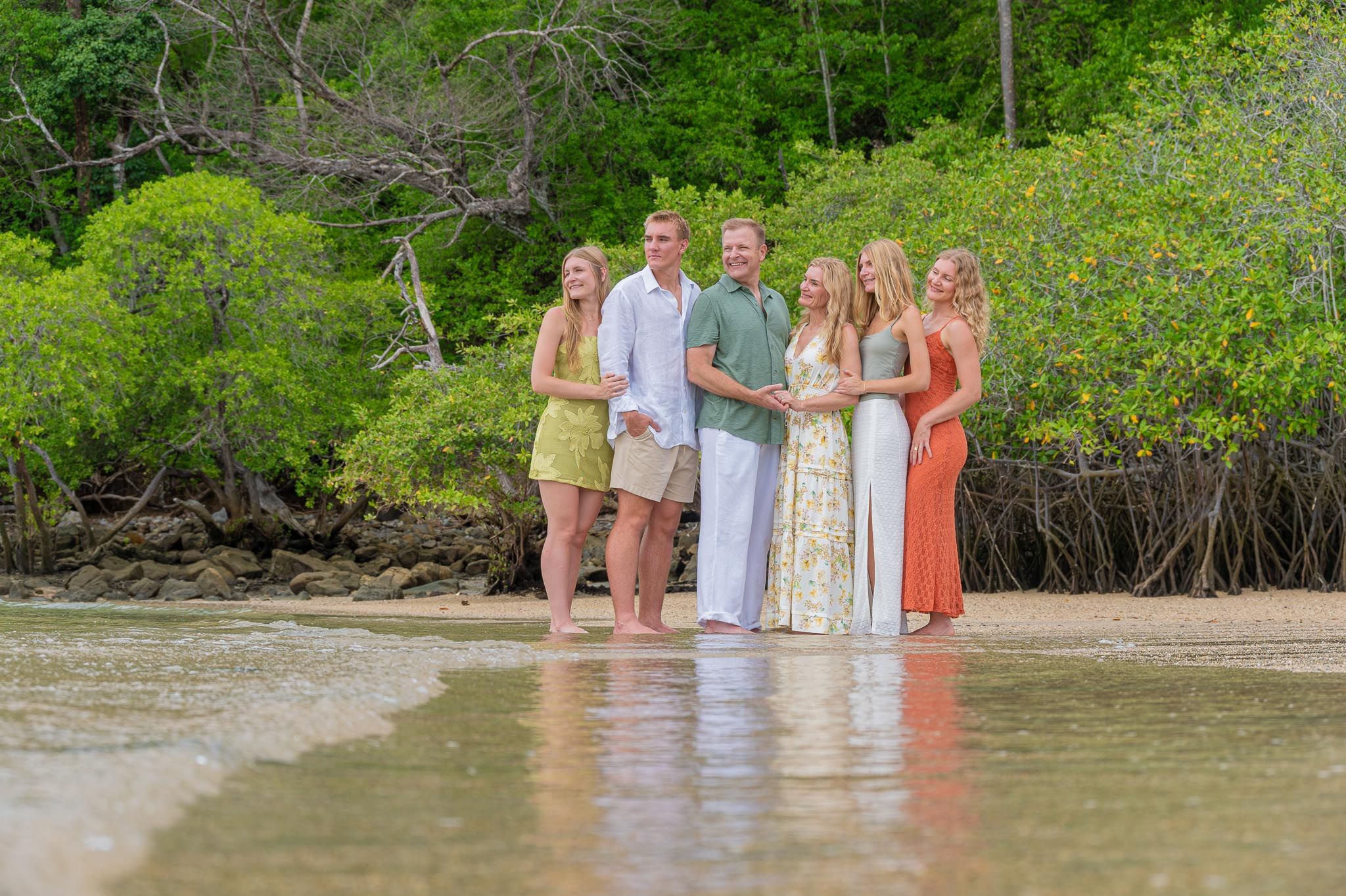 Family of six posing together on a tropical beach with lush green mangroves in the background.