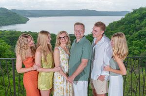 Family of six posing on a balcony overlooking a lush green valley and lake on a cloudy day.