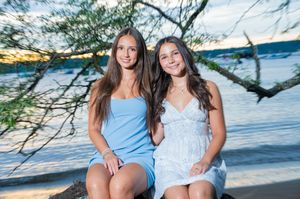 Two young women smiling together on a lakeside log at sunset, one in blue dress, one in white dress.