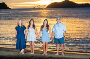 Family of four holding hands on a beach at golden sunset, wearing coordinating blue outfits with ocean and islands behind.
