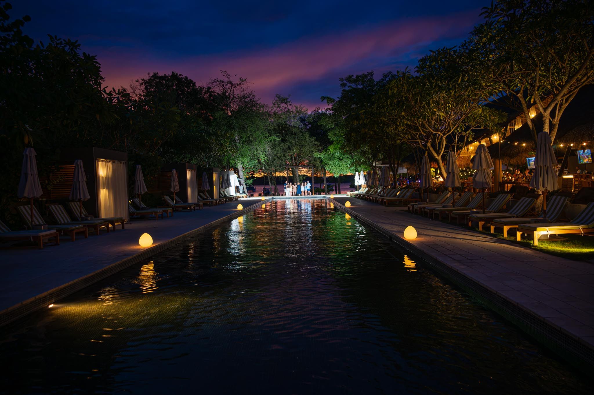 Illuminated resort pool at night with glowing lights reflecting on water, surrounded by tropical trees and lounge chairs.