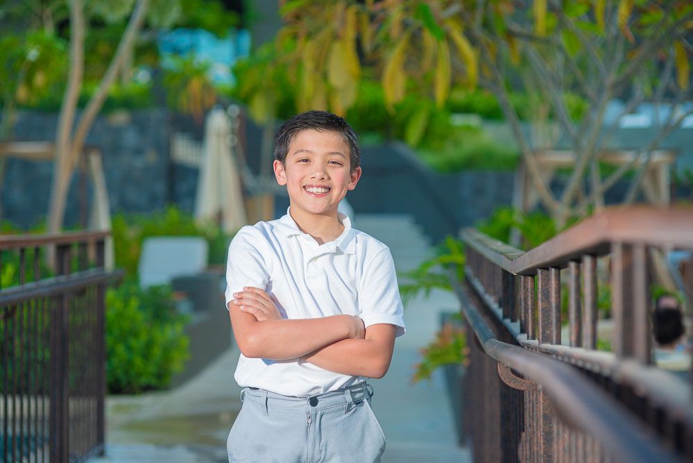 Smiling boy with arms crossed standing on a wooden bridge surrounded by tropical greenery.