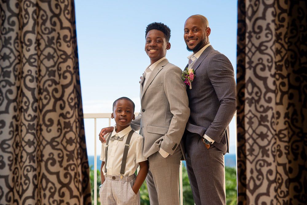 Three well-dressed males in suits smiling together, posing between decorative curtains at a formal event.
