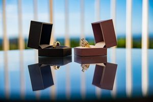 Two elegant gold rings displayed in open jewelry boxes on a reflective surface with a scenic blue sky background.