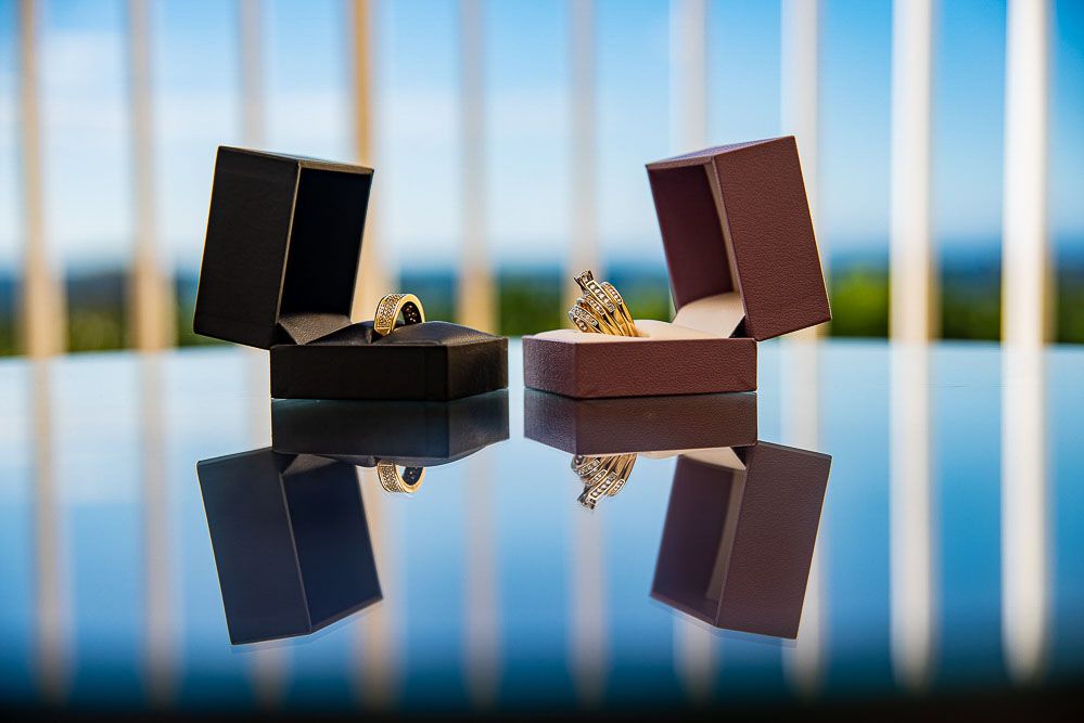 Two elegant gold rings displayed in open jewelry boxes on a reflective surface with a scenic blue sky background.