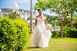 Bride in white wedding gown holding bouquet walks through lush green garden with tropical resort in background.