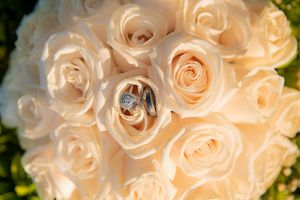Wedding rings resting on a bouquet of cream roses, featuring a diamond engagement ring and two gold bands.