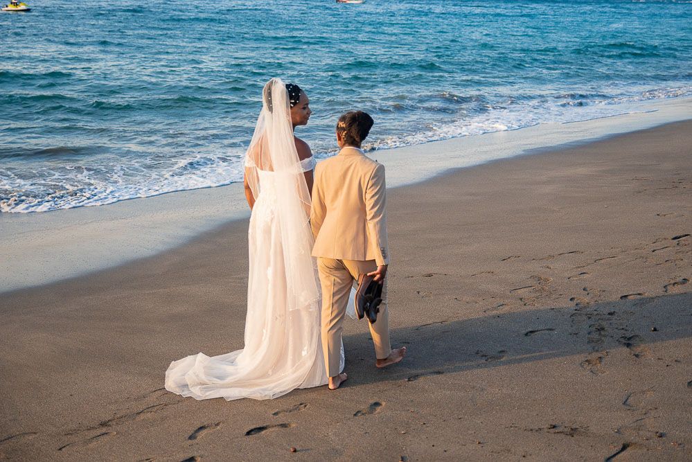 Newlywed couple in wedding attire walking hand in hand along a sandy beach with ocean waves.