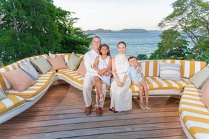 Family of four posing on a tropical deck with yellow striped sofas, ocean view, and lush greenery at sunset.