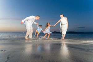 Family of four joyfully splashing in shallow ocean waves on a sandy beach at sunset.