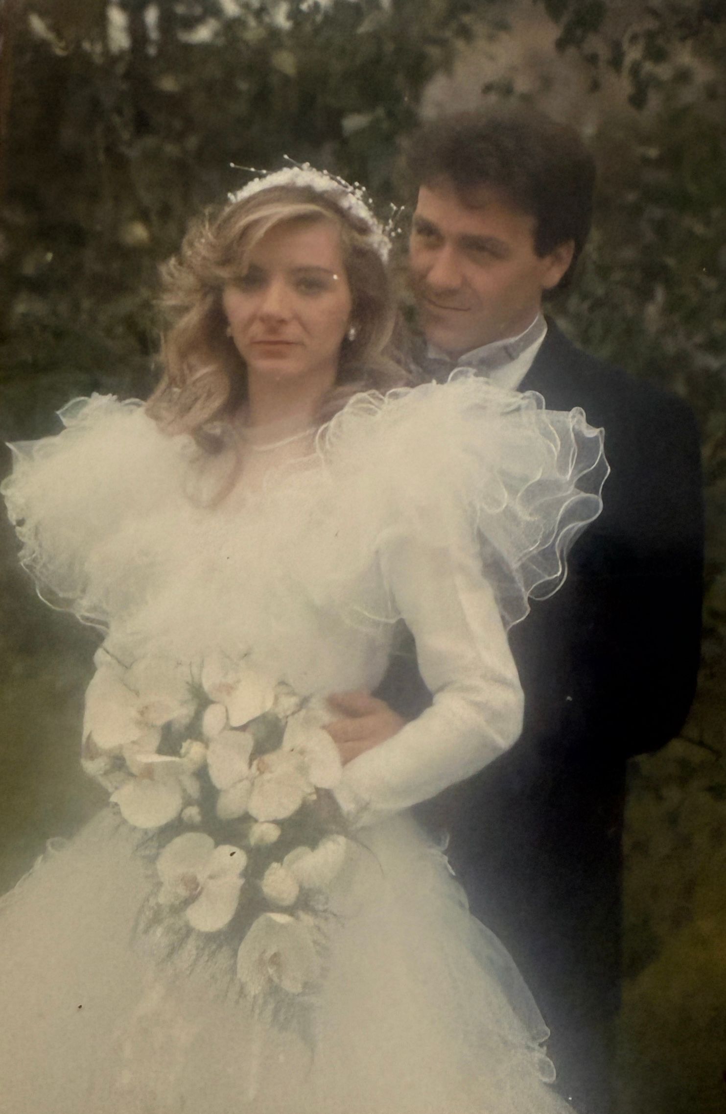 A vintage wedding photograph showing a couple in traditional attire with the bride wearing a white ruffled dress and holding roses.