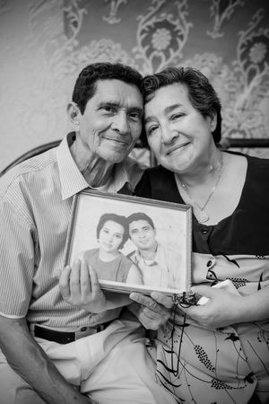 A couple sits together holding a framed vintage photograph of themselves from their younger years.