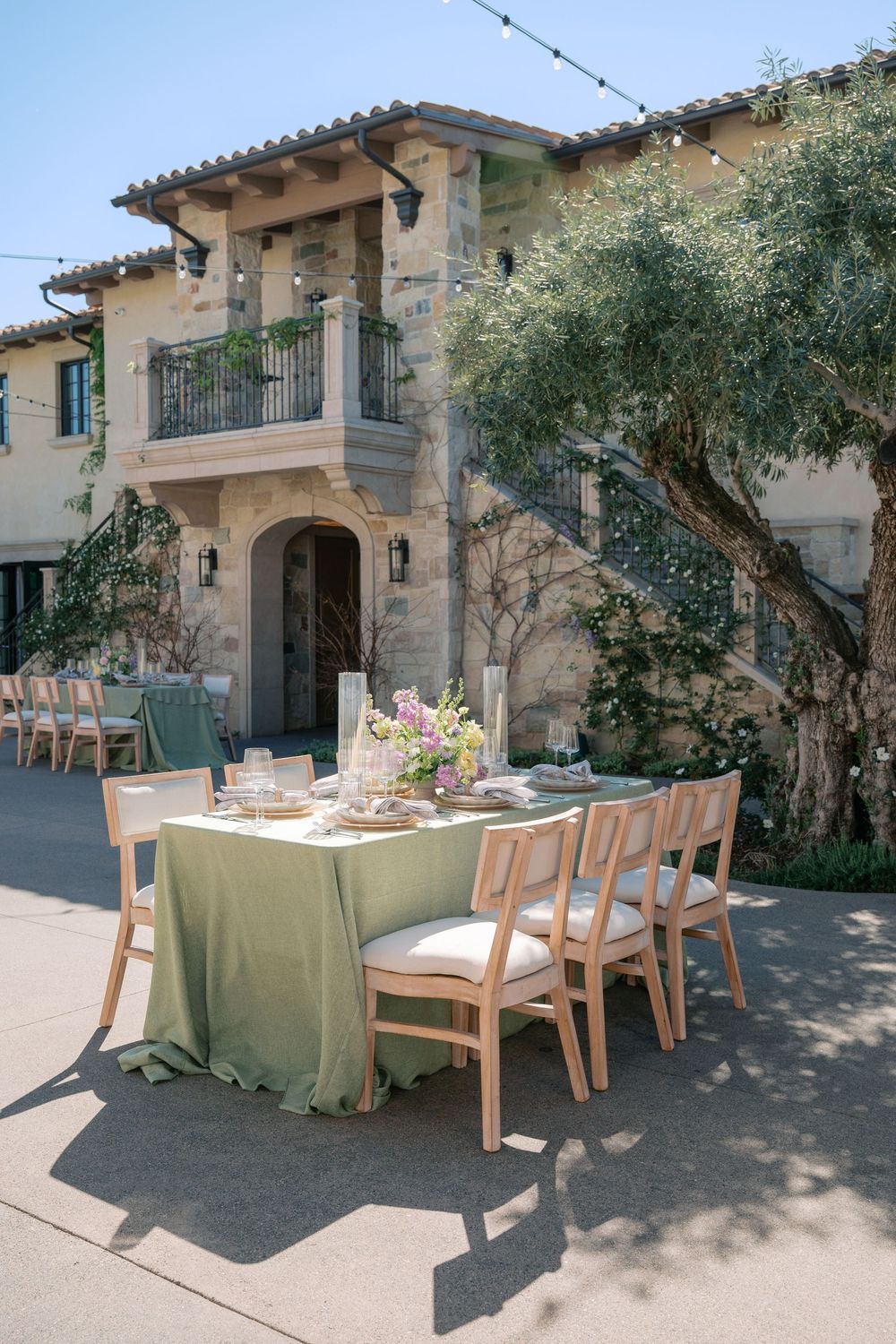 Elegant Mediterranean outdoor dining setup with wooden chairs and sage green tablecloths against a rustic stone villa.