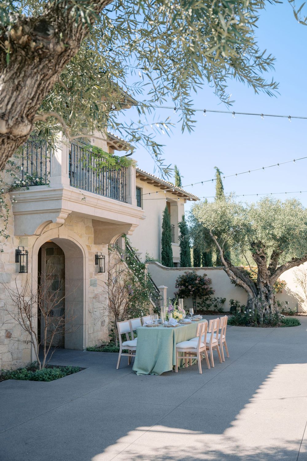 Mediterranean-style outdoor venue with cream stucco walls, olive trees, and string lights creating an elegant courtyard setting.