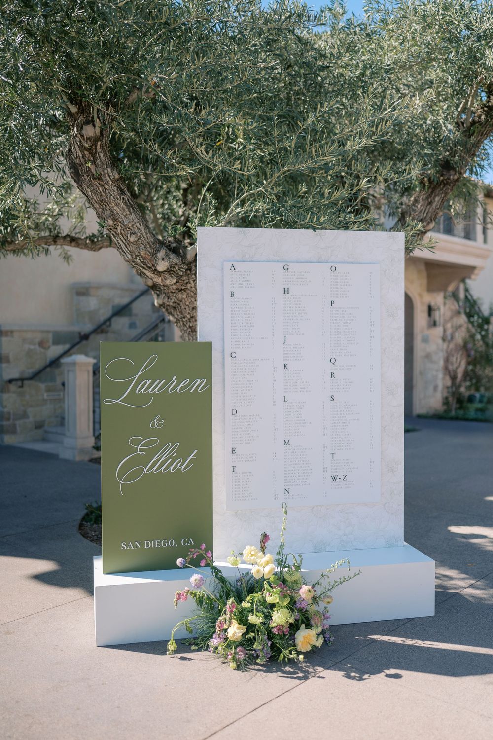 Wedding seating chart displays with olive green accents and floral decorations arranged outdoors.