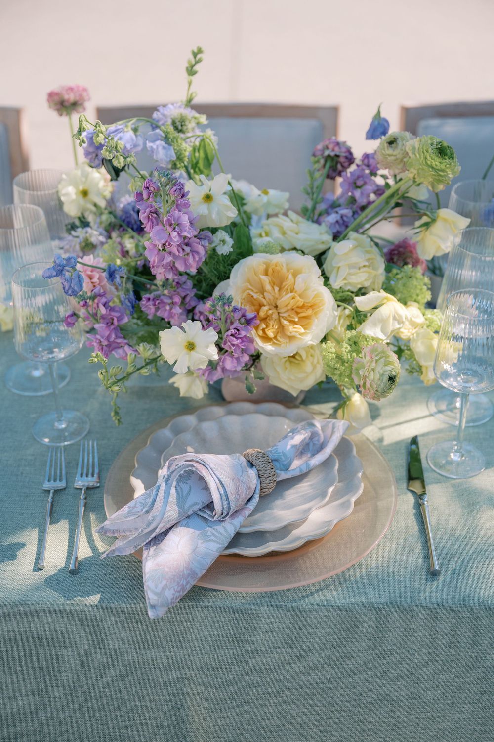 Elegant table setting with pastel floral centerpieces featuring lilac, yellow roses, and white blooms on a blue-gray tablecloth.