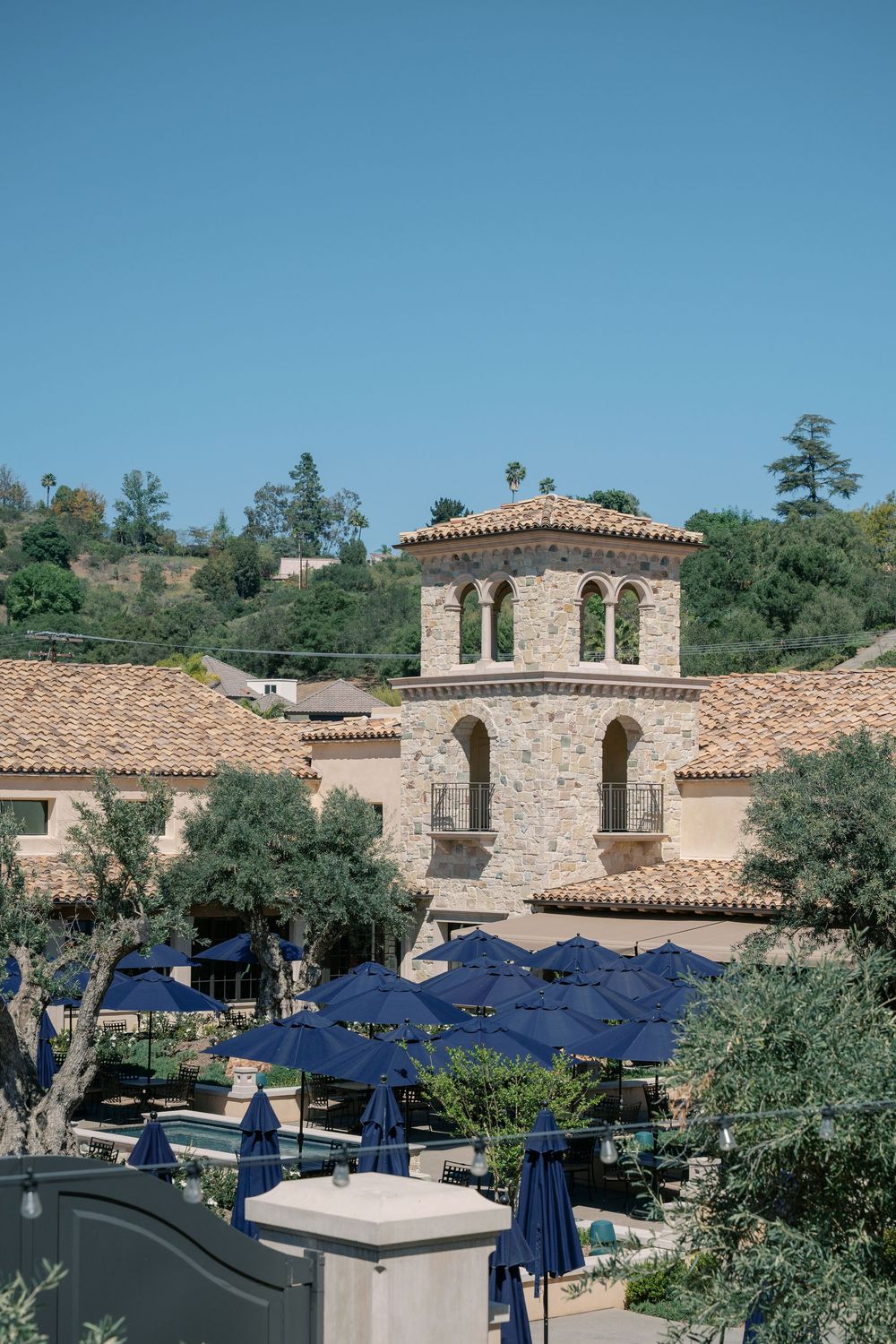 Mediterranean-style building exterior with terracotta roof tiles and blue patio umbrellas surrounded by lush greenery.