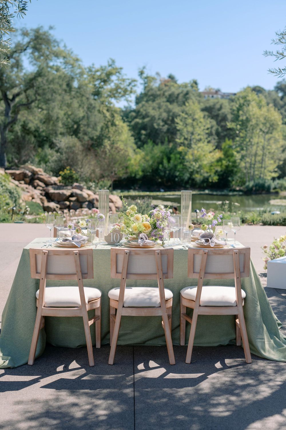 Elegant outdoor wedding reception table setup with sage green linens and light wood chairs along a paved pathway.