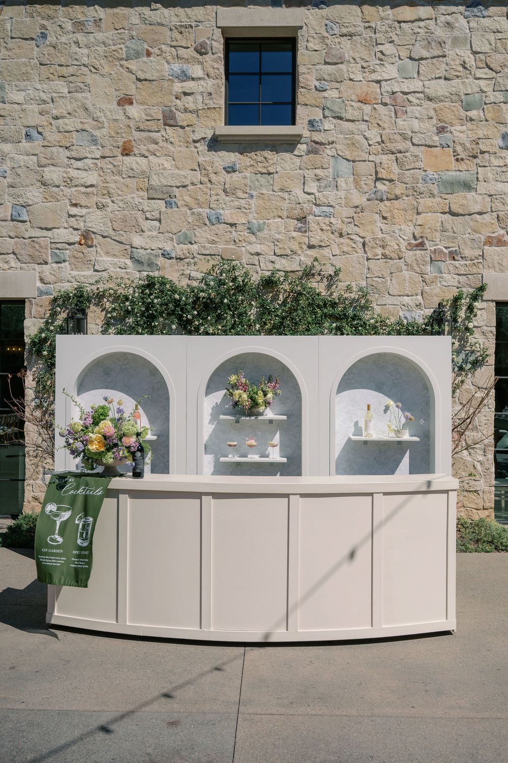 Elegant white display bar with three arched niches featuring floral arrangements and decor against a stone wall exterior.