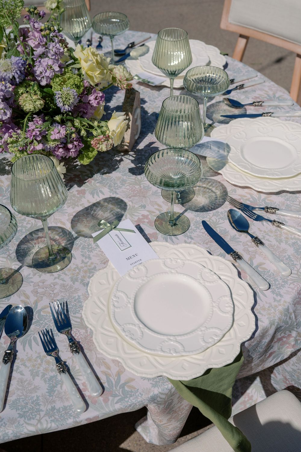 Elegant table setting with white embossed plates, green glassware, and floral arrangements on a patterned tablecloth.