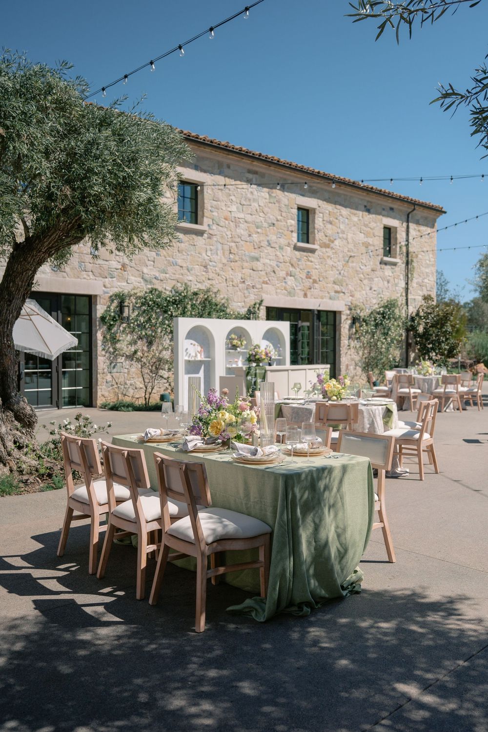 Elegant outdoor dining setup with long tables draped in sage green linens at a rustic Mediterranean stone villa terrace.