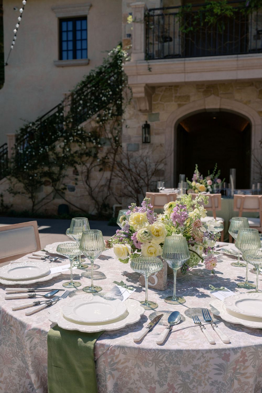 Elegant outdoor table setting with floral centerpieces, white plates, and glassware arranged on a textured tablecloth.