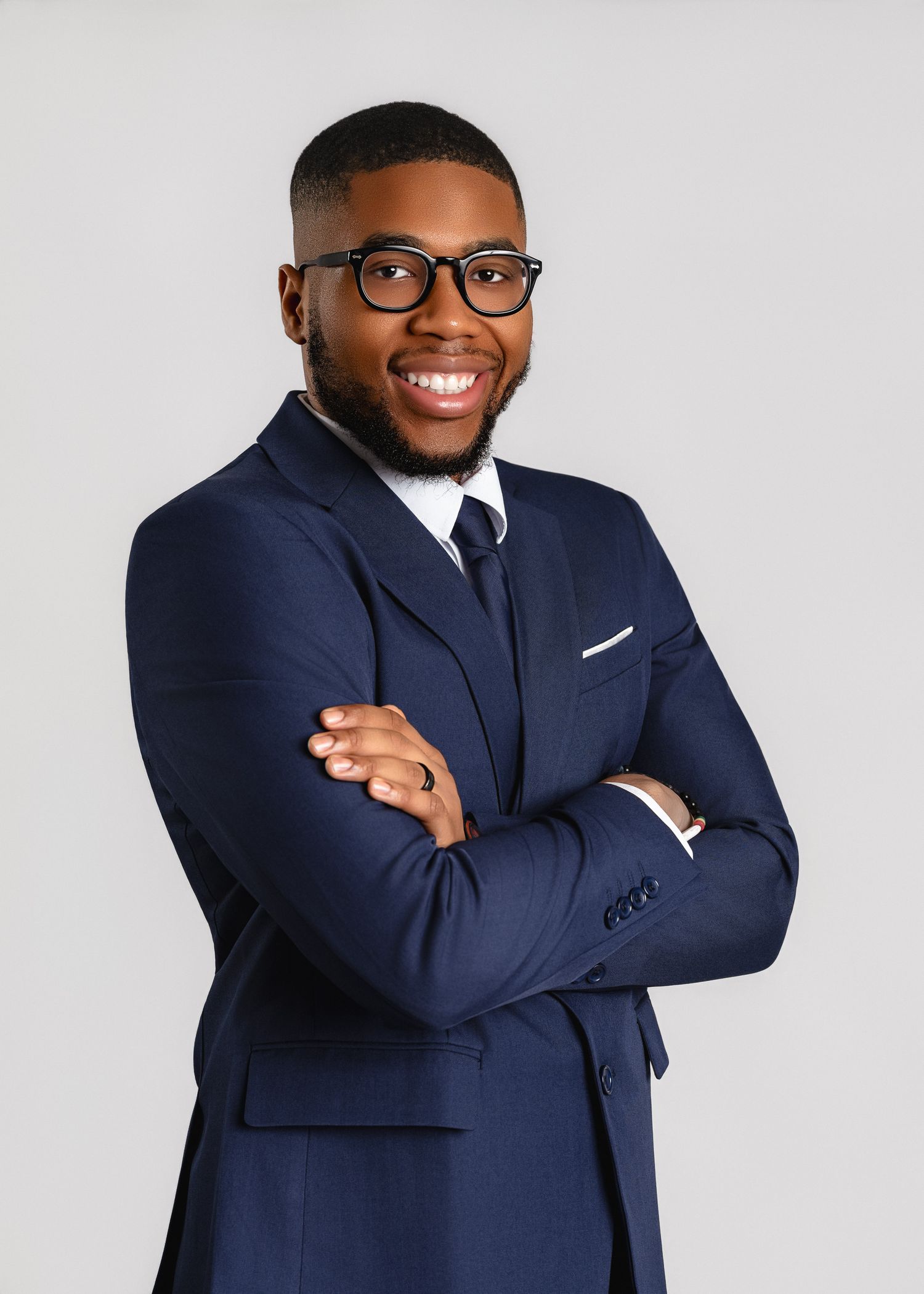 Professional wearing a navy blue suit, white dress shirt, tie and glasses poses with arms crossed against grey background.