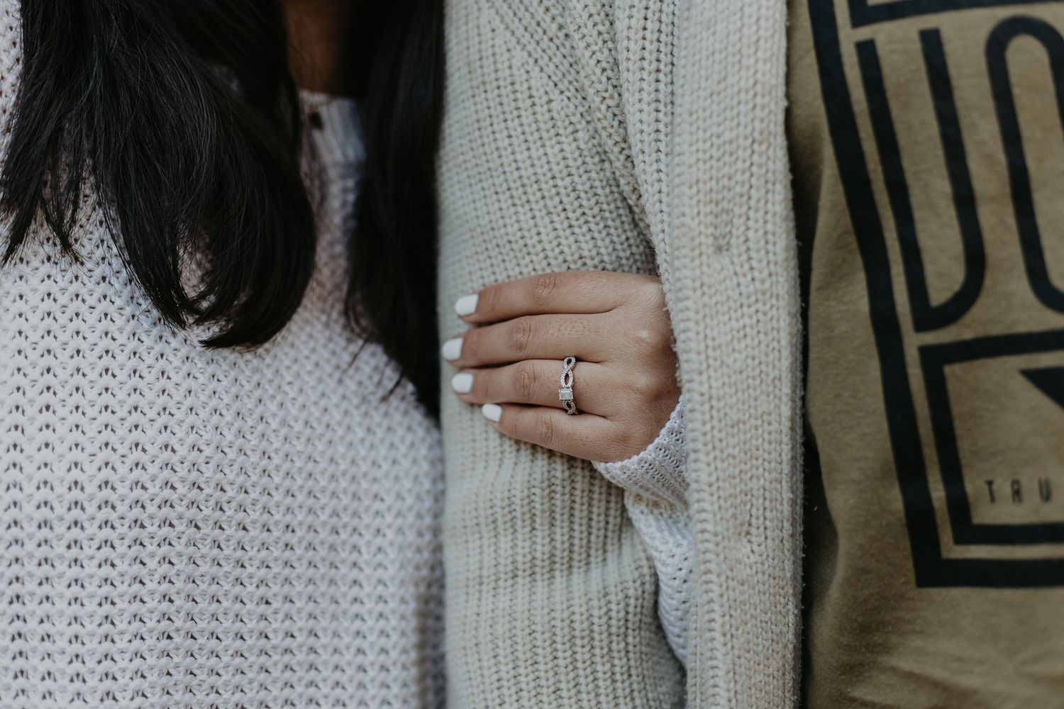 Close up of a woman in cozy cream sweater holding her fiance's arm to show her engagement ring.