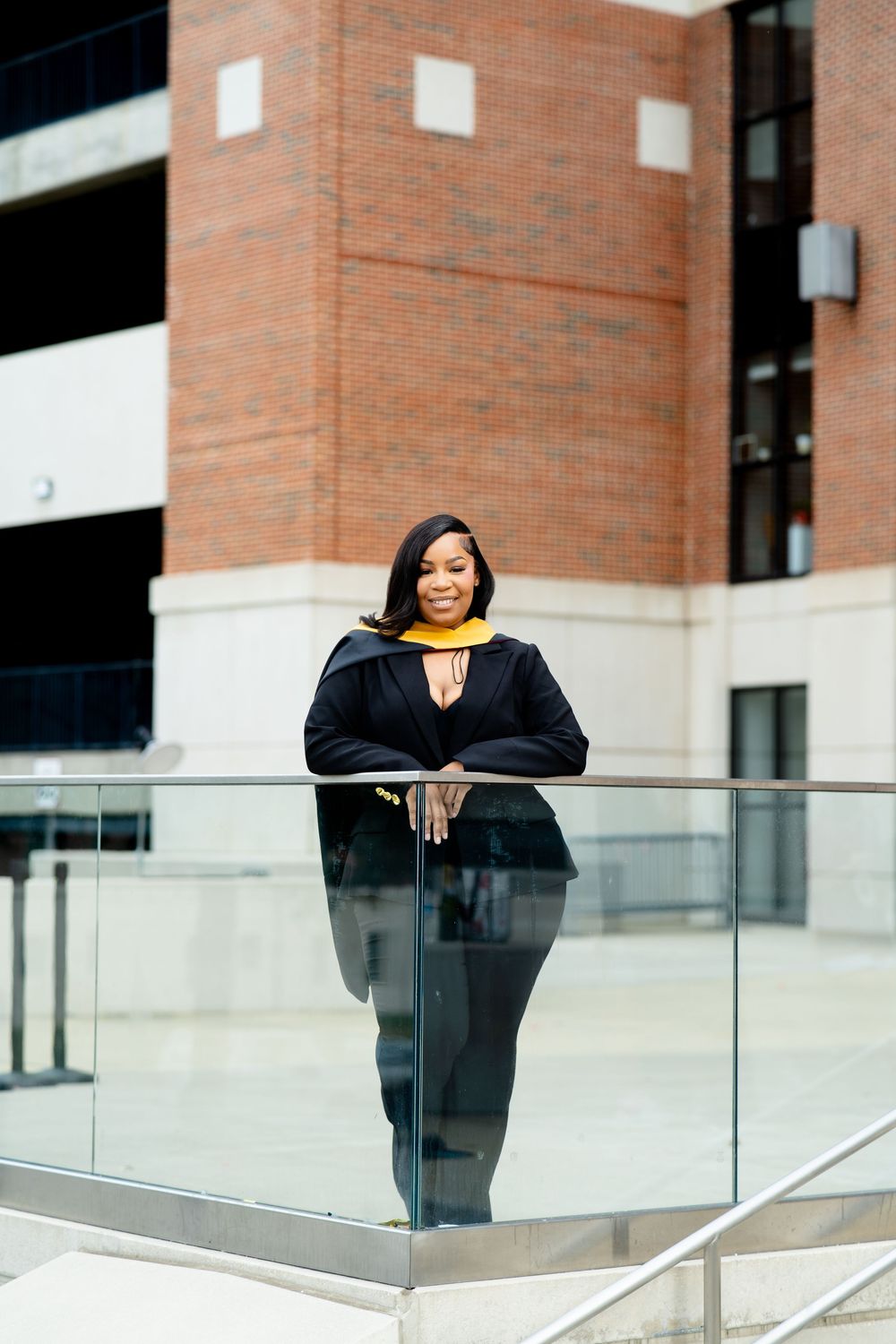 A smiling graduate in black gown and gold stole leans on a glass railing outside a brick building.