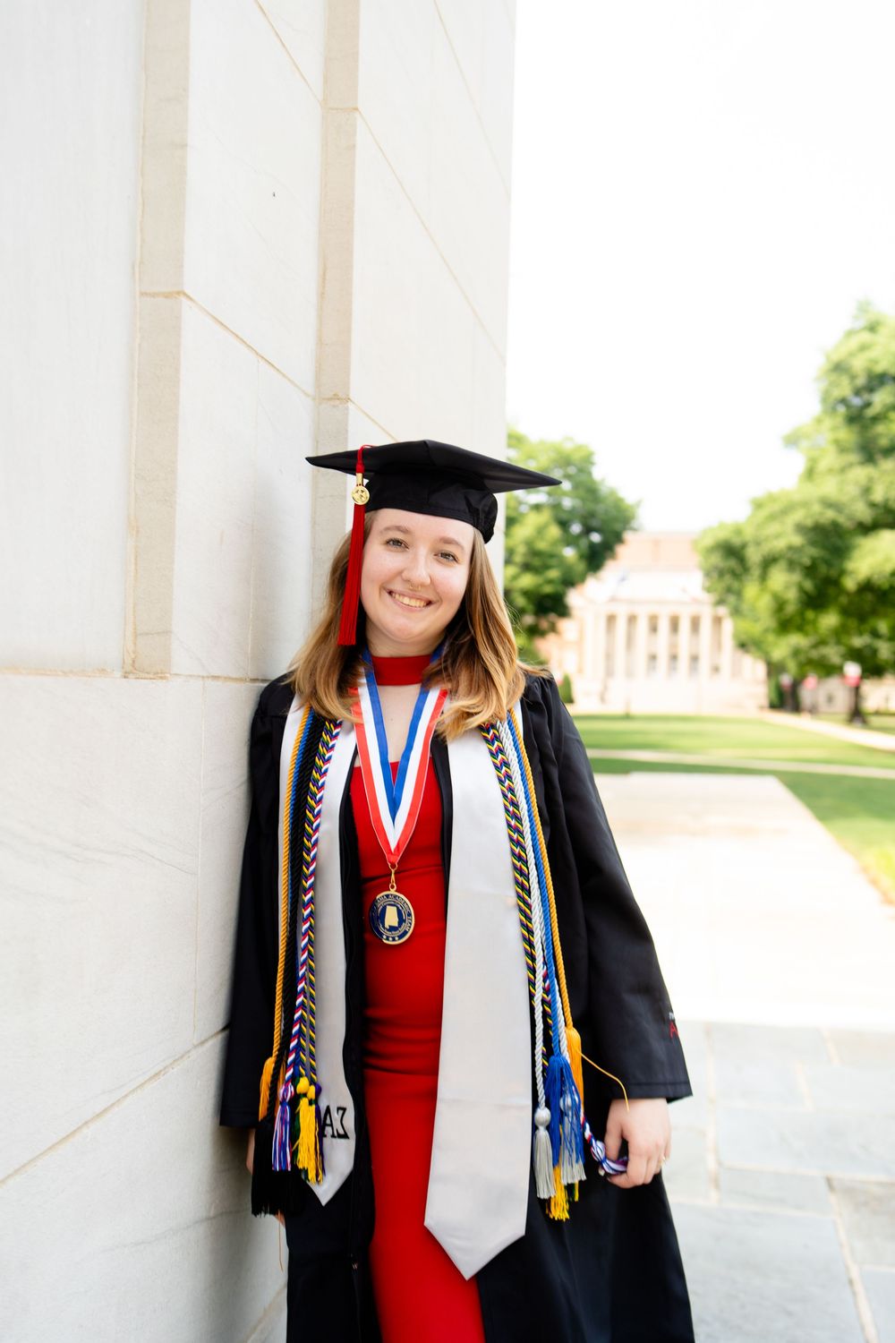 Smiling female graduate in black cap and gown with red dress, honor cords and medal, posing by a white column.