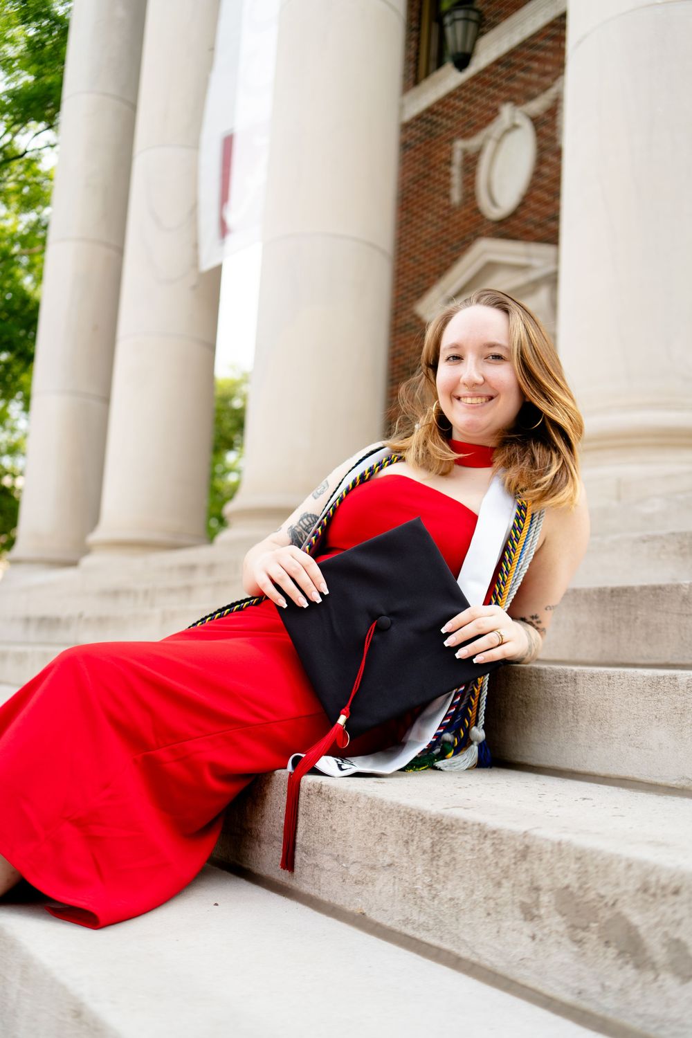 Smiling graduate in red dress holding black cap sits on stone steps near columns at university campus.
