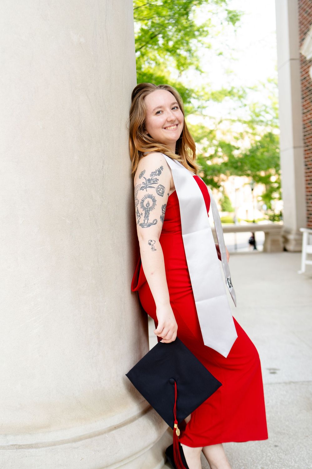 Smiling graduate in red dress with white sash holds cap, leaning against column with floral arm tattoo visible.