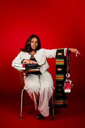 Young Black woman in white tweed suit sits at desk with books and a Black Girl Magic kente stole on red background.