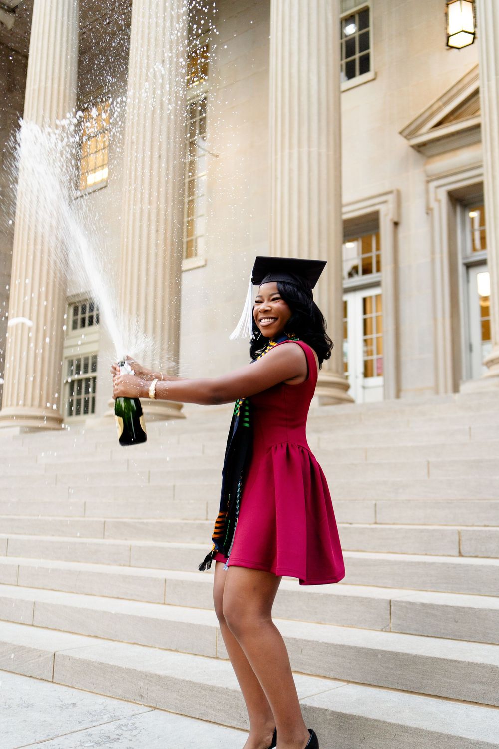 Young woman in red dress and graduation cap celebrates on university steps, spraying champagne bottle.