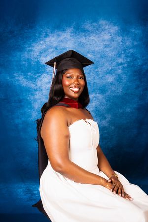 Smiling graduate in white strapless dress and black mortarboard cap poses against blue studio background.