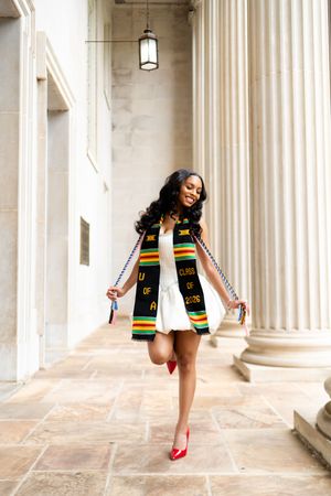 A joyful graduate poses between columns wearing a white dress, red heels, and a colorful kente cloth stole reading Class of 2016.