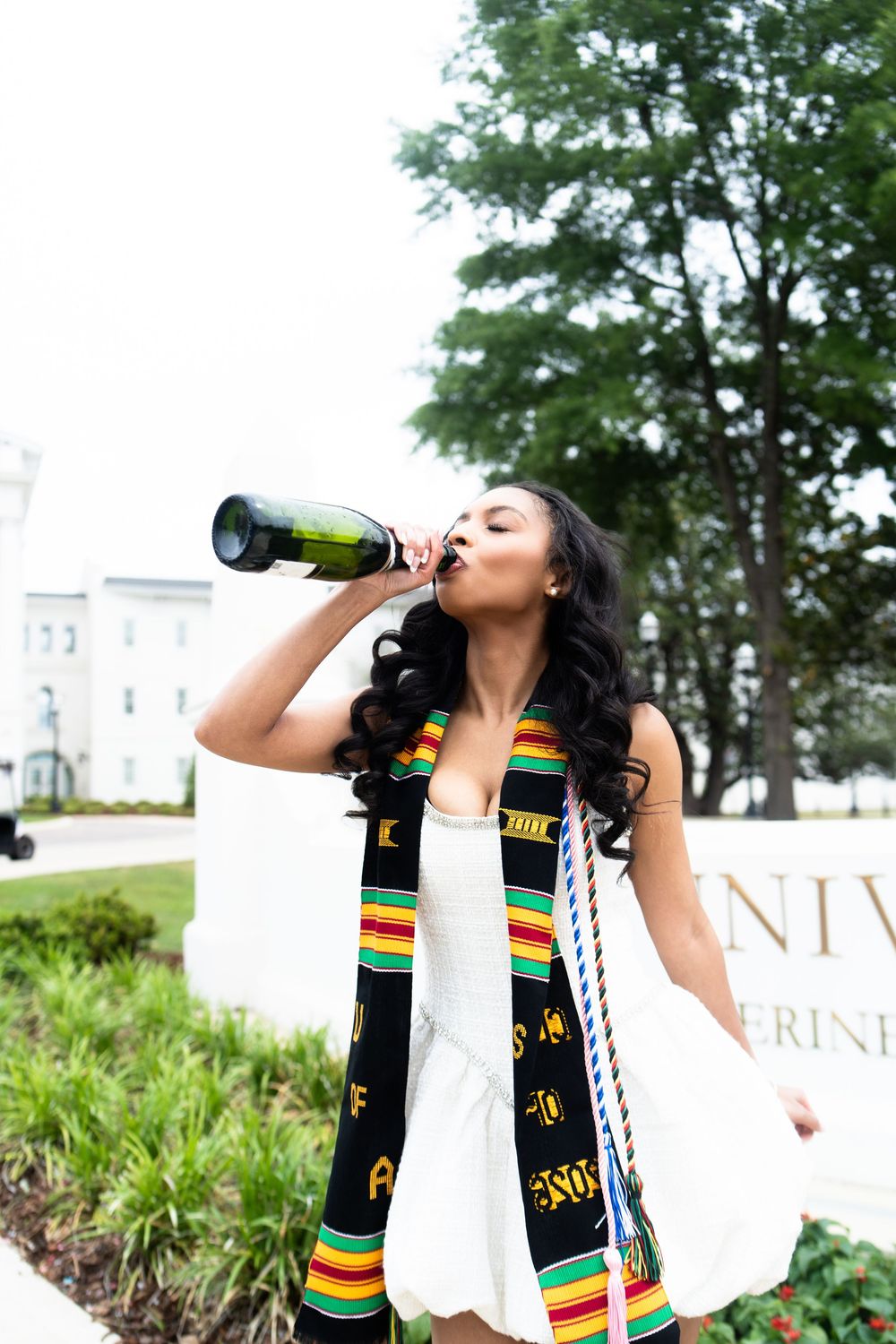 Graduate in white dress with kente stole and honor cords celebrates outdoors with champagne bottle.
