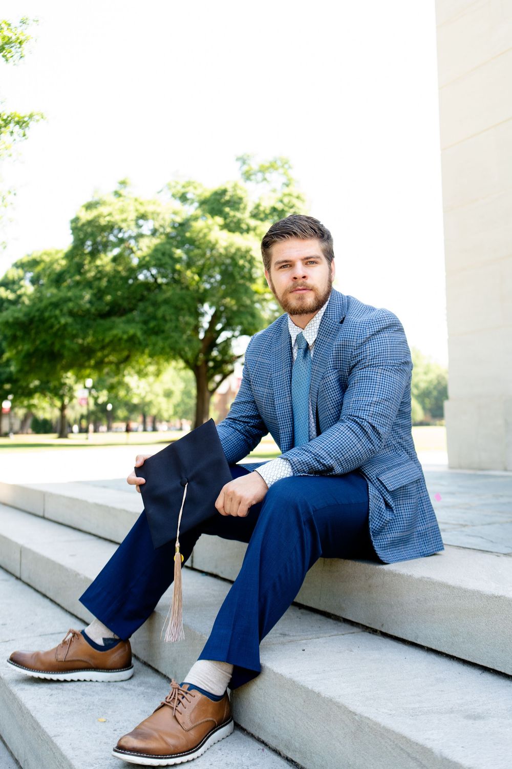 Young man in plaid blazer and blue tie sits on steps holding graduation cap outdoors.