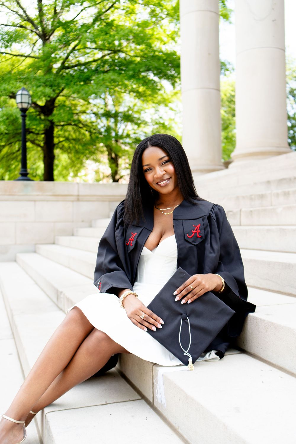 Smiling graduate in black University of Alabama gown sits on stone steps holding cap, wearing white dress outdoors.