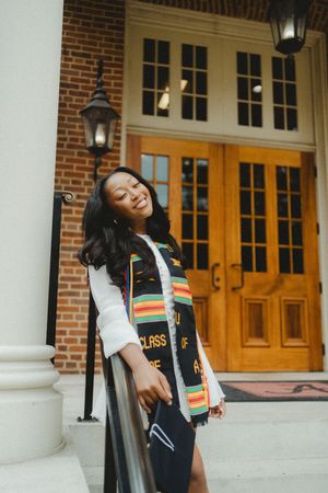 Graduate in cap and gown with colorful stole poses on steps of brick building with wooden doors and columns.