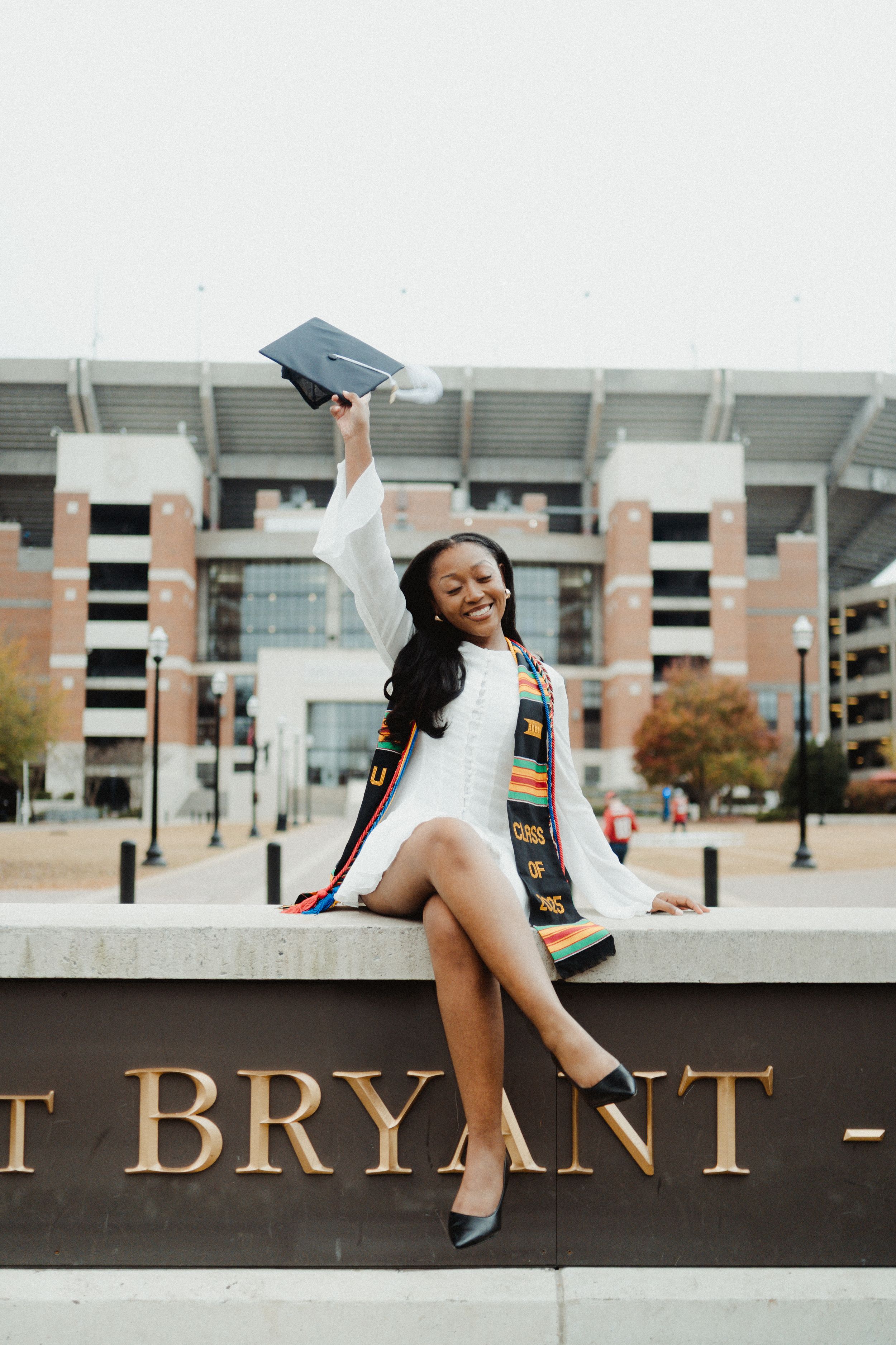 Graduate in academic regalia celebrates achievement with joyful poses on campus wall at Bryant University.