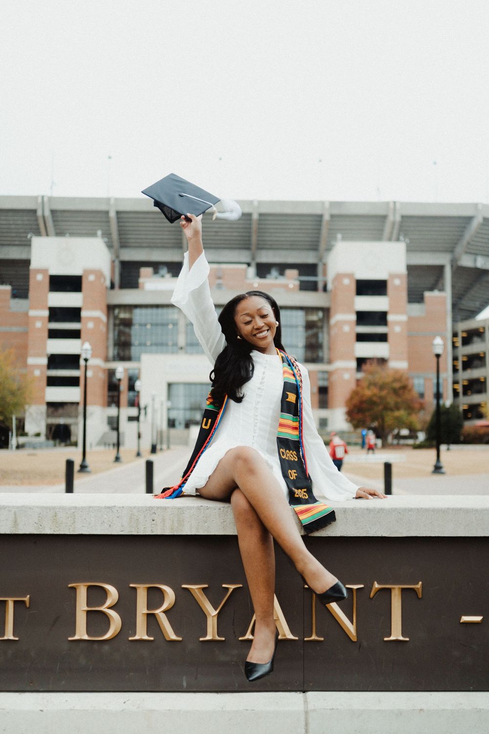 Graduate in academic regalia celebrates achievement with joyful poses on campus wall at Bryant University.