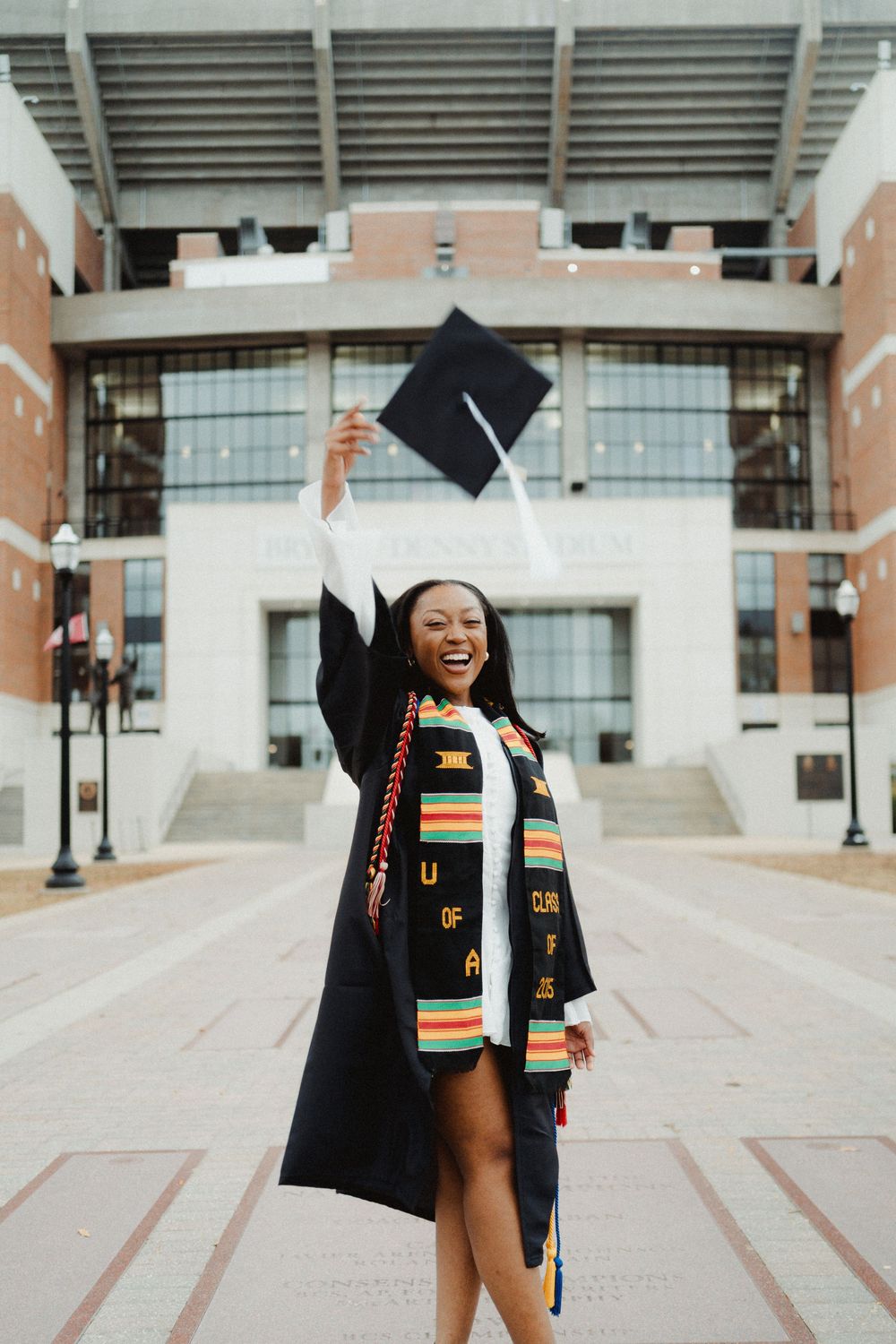 Graduate in cap and gown celebrating with raised arms in front of university building wearing colorful stole.